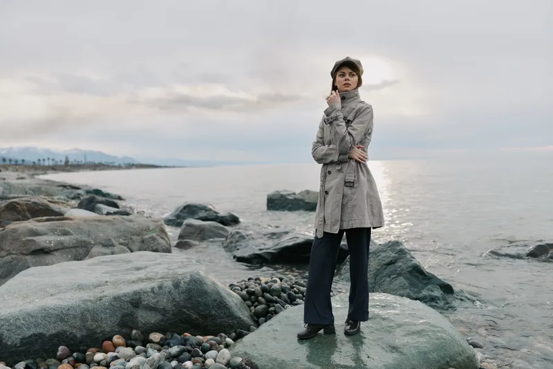 Young woman in long coat and hat stands on rocky beach near calm sea under cloudy sky. Casual fashion style in natural outdoor setting with soft daylight and muted colors.