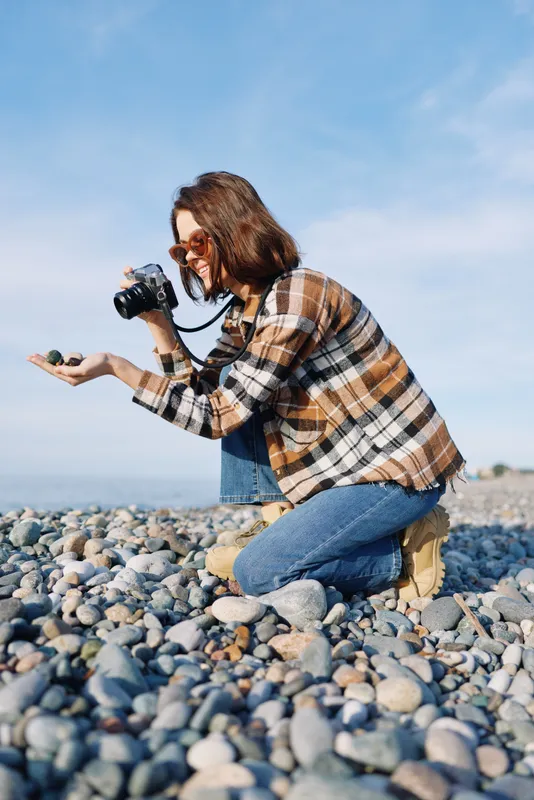 Photographer woman kneeling on a rocky beach, capturing with a compact camera, plaid shirt and denim jeans, clear blue sky, focused expression, outdoor creative moment by the coast.