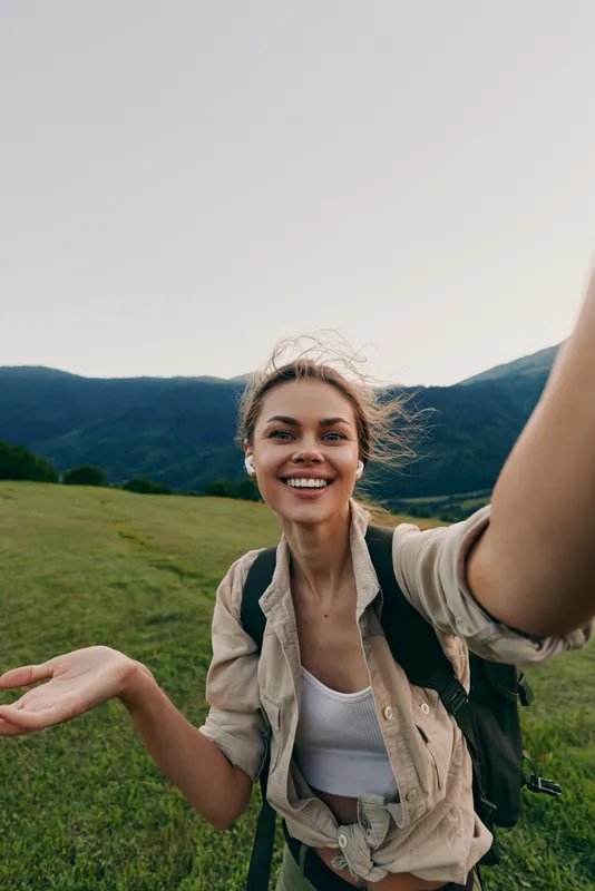 Smiling woman with backpack taking a selfie in an open meadow near mountains. Outdoor lifestyle, nature exploration, and travel mood. Bright natural light creates a warm vibe.