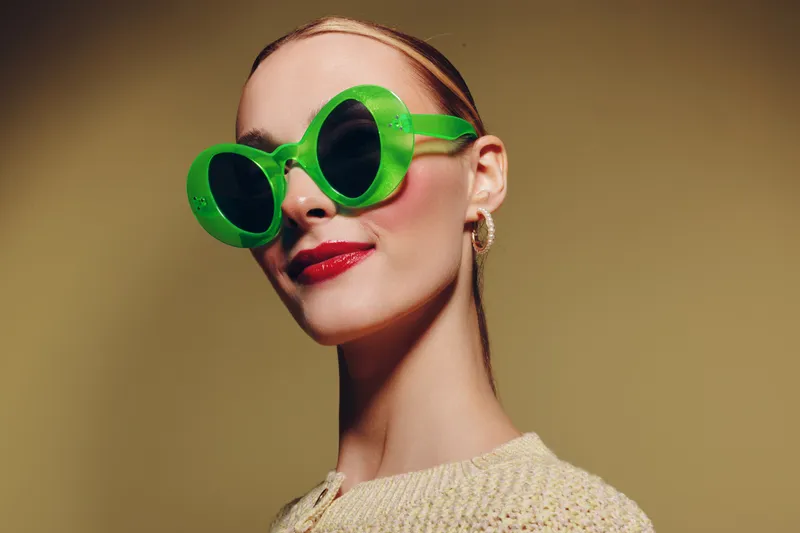 Stylish woman wearing large green sunglasses and red lipstick poses confidently against beige background. Concept of fashion, beauty, accessories, and modern style portrait.