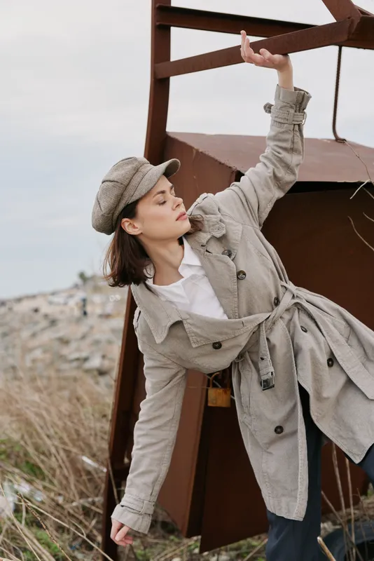 Young woman in beige trench coat and matching cap poses outdoors near rusty metal structure and dry grass, expressing calm and thoughtful mood on cloudy day. Fashion and autumn style concept.