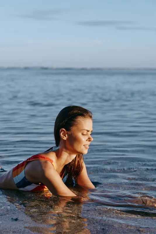Young woman in colorful swimsuit relaxing in shallow sea water during sunny day. Calm ocean background with clear sky. Concept of summer vacation and beach leisure.