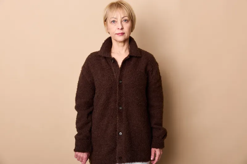 Elderly woman portrait, standing in beige studio background, dark coat and casual knitwear, calm mature expression, neutral warm light, copyspace for text, quiet