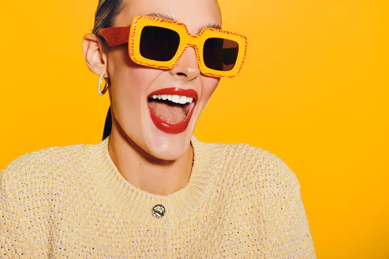 Happy woman wearing large orange sunglasses and a beige sweater laughs joyfully against a bright yellow background. Concept of summer style, fashion, and positive emotions.