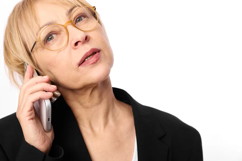 Woman in business suit with glasses holding phone to ear, studio portrait of blonde professional, direct gaze, calm expression, customer call, clean white background