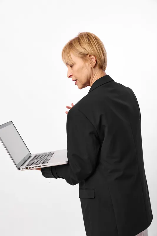Stressed businesswoman in black blazer works on laptop in office setting, anxiety shows on her face, she types and glances down, work pressure concept with technology and mental strain