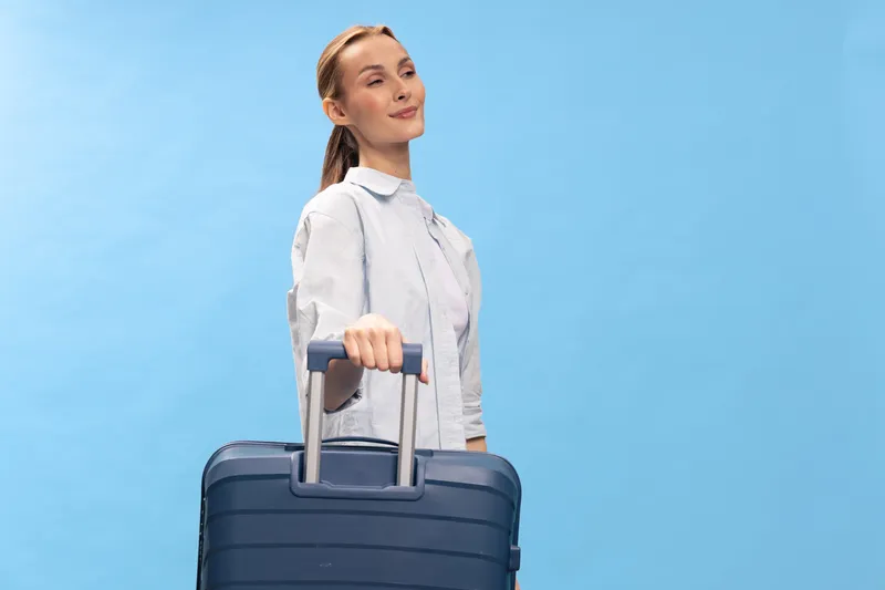 Young woman in casual shirt holding blue suitcase on blue background. Concept of travel, vacation, and summer trip with positive mood and light studio lighting.