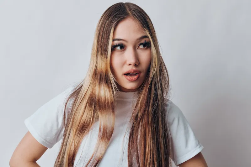 Young woman with long straight hair wearing a white t shirt looking surprised or curious on gray background with casual pose and natural expression.