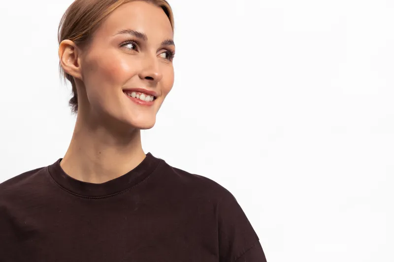 Smiling woman with light skin and blonde hair looks to the side wearing a dark brown shirt on a white background. Concept of happiness, casual style, and positive emotion.