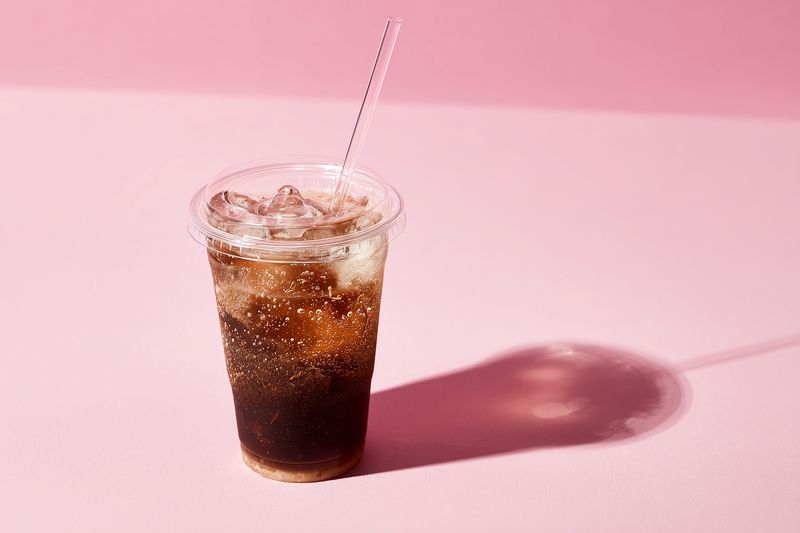 Cold fizzy cola in a clear plastic cup with ice and a straw on a pastel pink background, casting a soft shadow; refreshing beverage idea for summer, drink and lifestyle concepts.