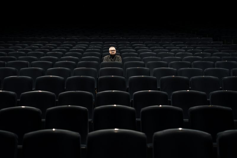 A lone person sits in the center of an expansive dark auditorium surrounded by countless empty seats, evoking solitude, contemplation, dramatic emptiness and quiet atmosphere.