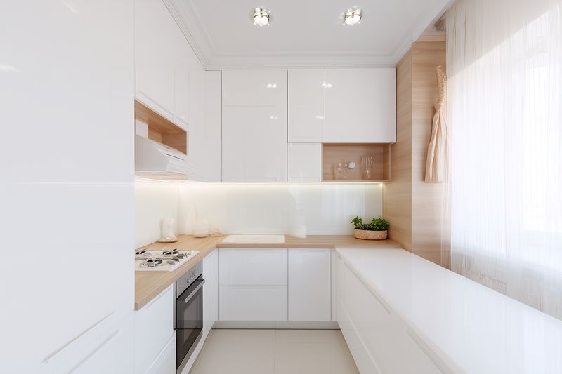 Bright modern white kitchen interior with sleek glossy cabinets, wood countertops, integrated appliances, soft undercabinet lighting and large window creating a clean minimalist space.