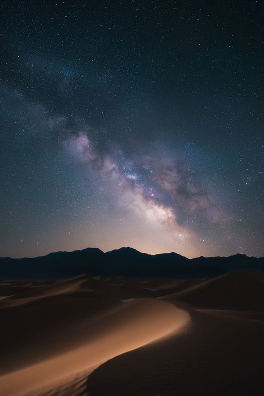 Long exposure night sky over desert dunes and silhouetted mountains, showing a bright galaxy core and countless stars above rippled sand under a clear cosmic panorama.