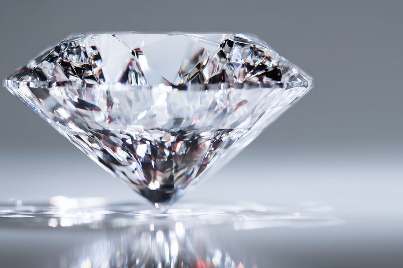 Close-up of a brilliant cut diamond on a reflective surface, showcasing sharp facets, light dispersion and sparkling highlights in a minimalist studio composition with soft background.