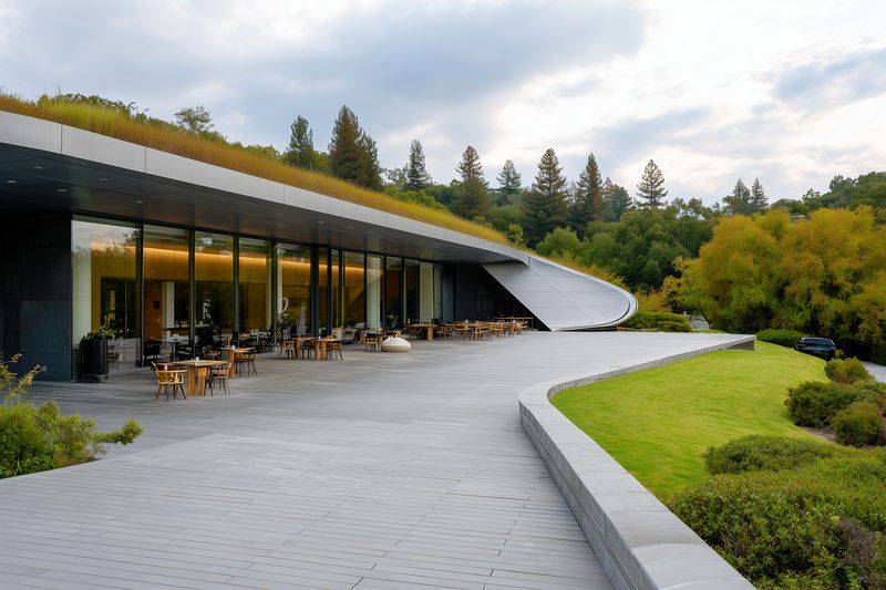 Modern building with glass facade and spacious outdoor terrace featuring tables and chairs, a green roof and manicured lawn, surrounded by trees and natural hillside under cloudy sky.