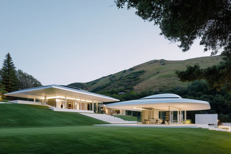 Modern minimalist glass house with illuminated interiors set on a manicured lawn, featuring low sloping roofs and open pavilion design against rolling hillside at dusk.