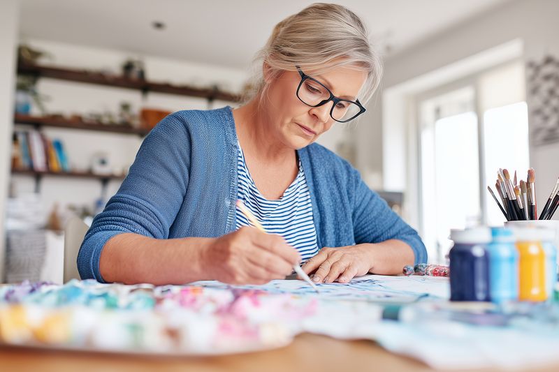 Senior woman painting at home studio, focused on watercolor artwork at a cluttered table with brushes, palette and jars, expressing creativity and a peaceful artistic hobby in natural light.
