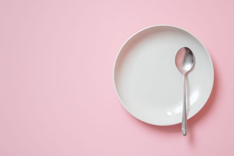 Top view of a white ceramic plate and a stainless steel spoon on a pale pink background, minimalist flatlay composition suggesting empty tableware and a simple dining concept.