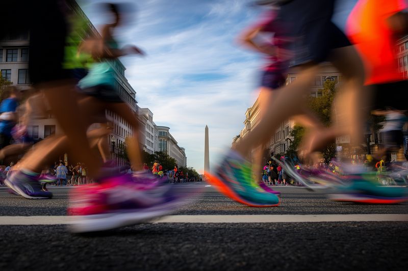 Low-angle motion blur of colorful runners crossing a busy urban street in a road race, capturing dynamic legs and sneakers, energetic crowd, and vivid sense of speed and endurance.
