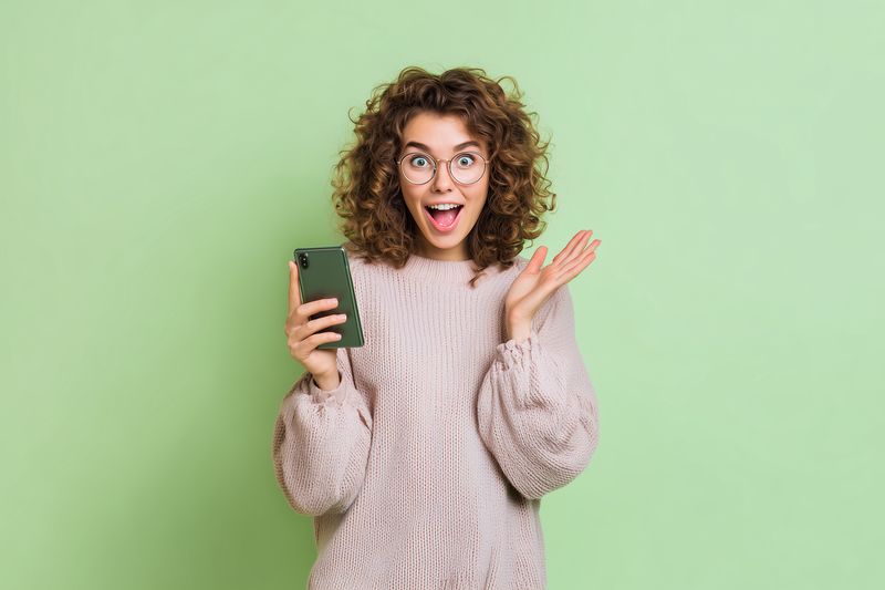 Surprised young woman with curly hair and glasses holds a smartphone while expressing joyful astonishment against a soft green background, conveying excitement and modern communication.