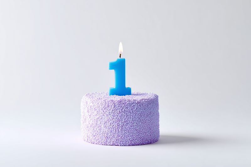 Close-up of a small purple textured cake adorned with a lit blue number one candle, presented in a minimalist studio setup on a neutral background to evoke a first birthday celebration.