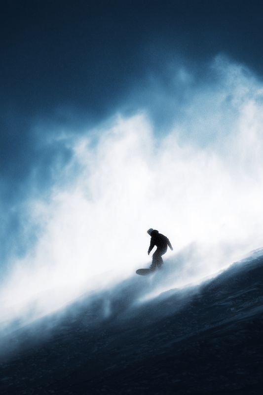 Silhouetted snowboarder carving down a steep snowy slope through windblown powder under dramatic low light, motion and spray capturing adrenaline and raw winter mountain energy.