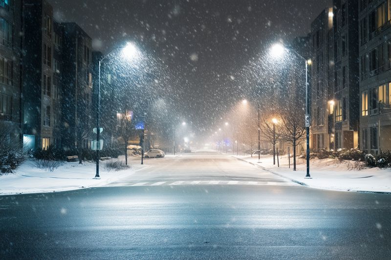 Quiet urban street at night during heavy snowfall, illuminated by streetlights reflecting on wet pavement, with modern buildings flanking the road and fresh snow accumulating.
