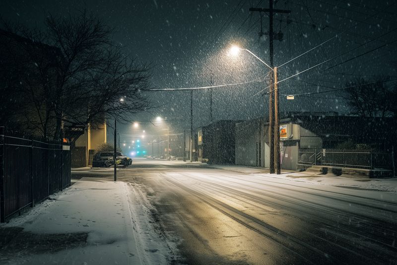 Snow falls on a quiet urban street at night, illuminated by glowing streetlights and storefronts. Wet pavement reflects warm light, creating a moody, atmospheric winter scene.