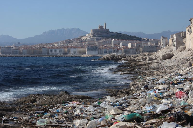 Coastal urban shoreline strewn with plastic and household debris along rocky beach, contrasting distant cityscape under clear sky, illustrating severe marine pollution and neglect.