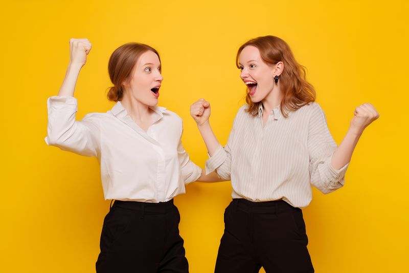 Two young women enthusiastically celebrating with raised fists and joyful expressions against a vivid yellow backdrop, capturing energetic emotion, victory, camaraderie and friendly connection.