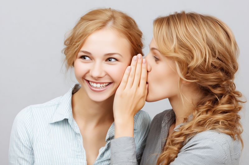 Two young women share a playful secret as one whispers into the other's ear, both smiling in a bright studio portrait that conveys friendship, intimacy, and joyful communication.