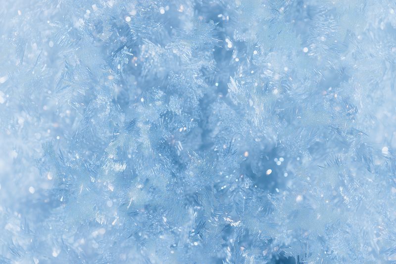 Close-up macro texture of fresh snow and ice crystals with soft blue tones, detailed frozen surface capturing glistening crystalline structure and serene winter abstract background.