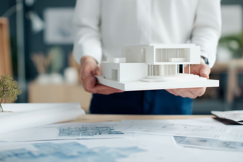 Person holding a white architectural model over detailed blueprints on a workspace, illustrating modern house design, architectural planning, scale modeling, construction concept and creative