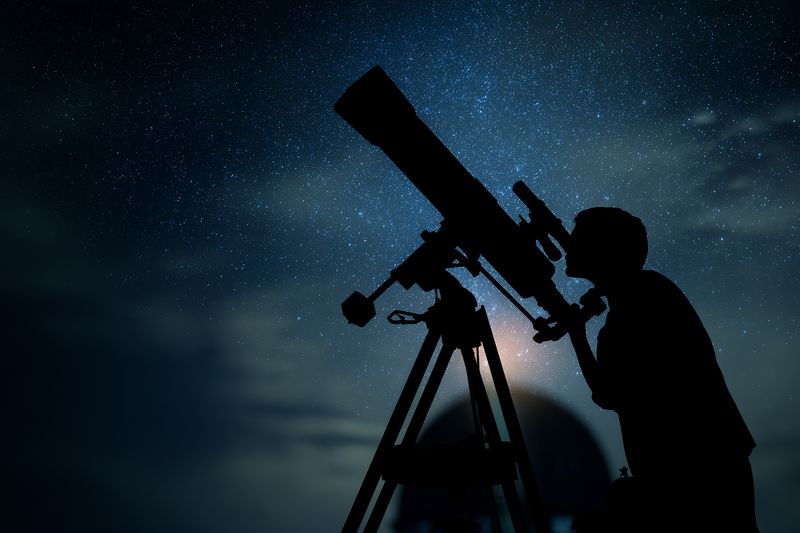 Silhouette of a person observing the night sky through a telescope under a starry clear sky, capturing astronomical hobby, exploration and wonder in a serene outdoor setting.