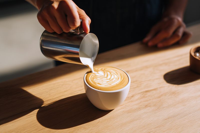 Barista pouring steamed milk into a ceramic cup to create latte art, close-up of hands crafting a heart pattern on espresso crema on a wooden counter in warm natural light.