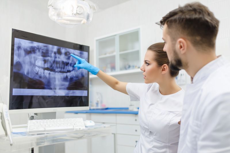 A female dentist wearing blue gloves points at a dental X-ray displayed on a monitor while explaining findings and treatment options to a male colleague in a bright clinic room.