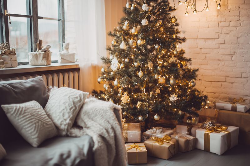 Cozy living room with a decorated Christmas tree illuminated by warm fairy lights, presents wrapped in kraft paper and gold ribbon, soft sofa with textured pillows and knit blanket.