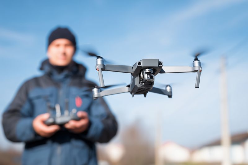 Compact drone hovering in sharp focus over a residential area while a remote pilot operates controls in the blurred background, illustrating aerial photography and hobby flying.