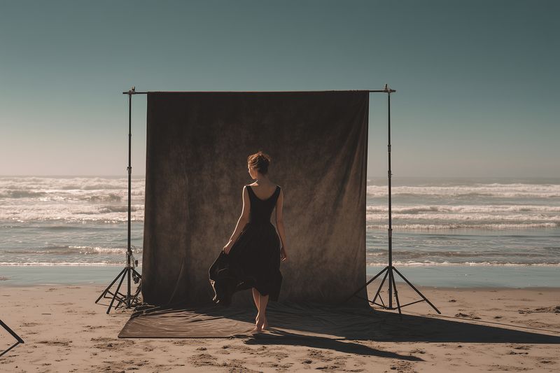 A lone woman in a flowing black dress stands barefoot on a sandy beach before a textured backdrop, sunlight casting long shadows in a minimalist coastal portrait setup.