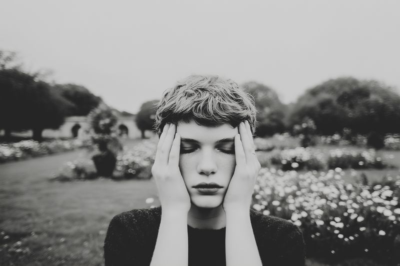 Black and white portrait of a contemplative young woman in a garden, hands pressed to her temples, moody vintage grain and soft shallow focus conveying introspective emotion.