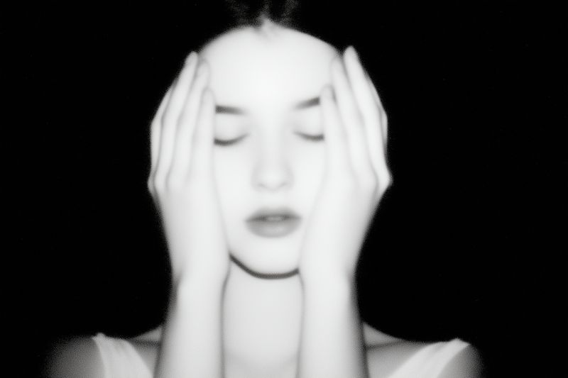 High-contrast grainy black and white portrait of a contemplative woman with closed eyes and hands pressed to her face, evoking introspection, melancholy and cinematic vintage mood.