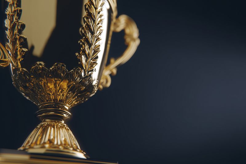 Close-up of an ornate golden trophy cup with intricate laurel detailing, symbolizing victory, achievement and excellence, photographed with shallow depth of field and moody lighting.