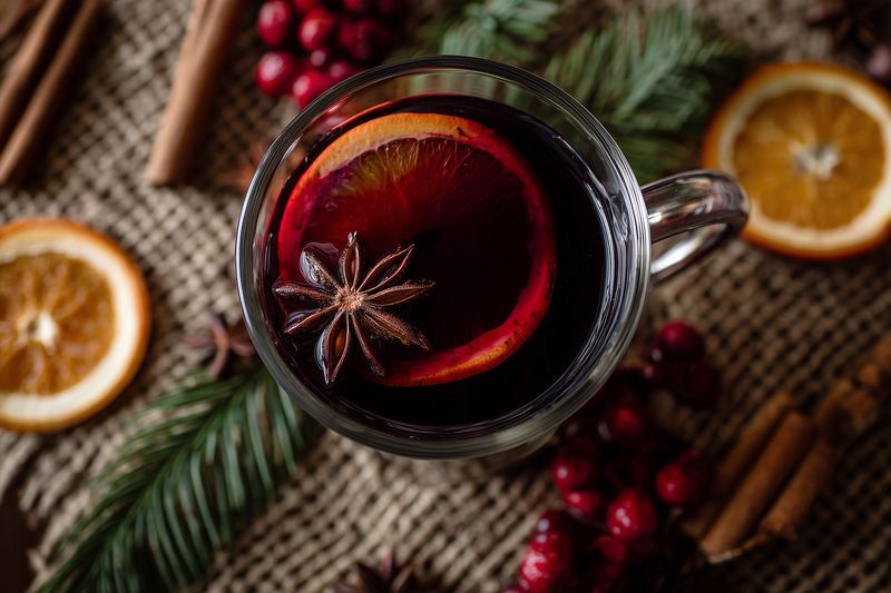 Close-up overhead of a warm spiced mulled wine in a clear glass mug with orange slice and star anise, arranged on a rustic cloth with cinnamon sticks, pine sprigs and red berries.