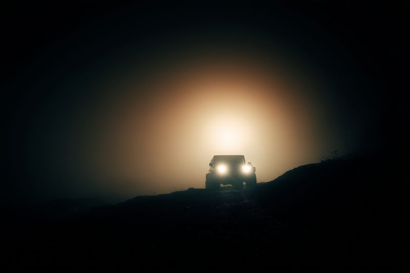 A solitary vehicle emerges from dense fog at night, headlights piercing the darkness as it travels along a remote road, creating a dramatic and mysterious silhouette.