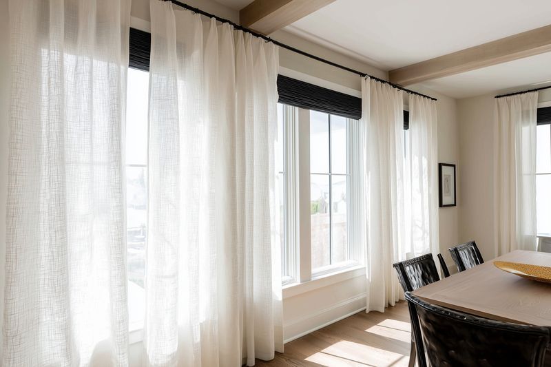 Bright dining room with sheer white curtains filtering sunlight through large windows, wooden beams overhead and a cozy table with chairs creating a warm modern farmhouse atmosphere.