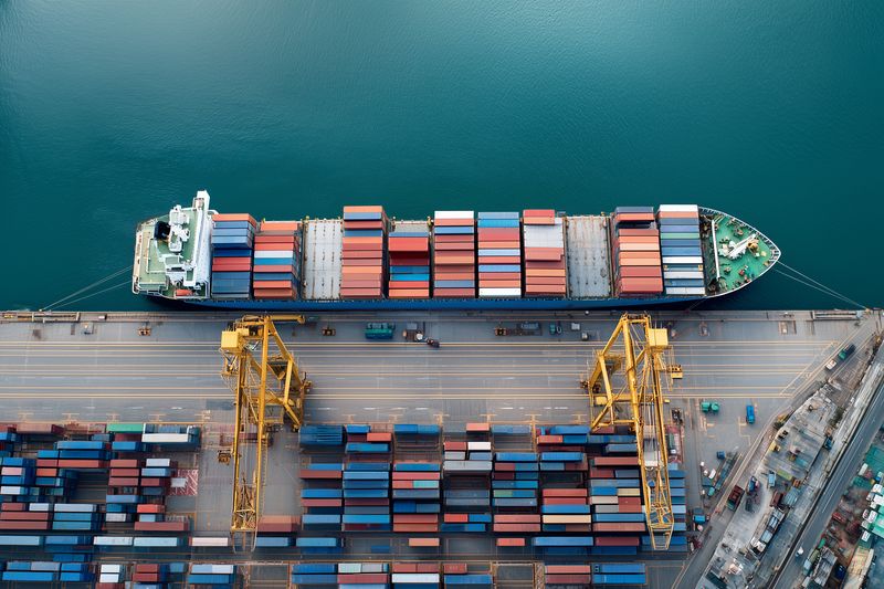 Aerial view of a large container ship docked at an industrial port terminal, yellow gantry cranes loading and unloading multicolored shipping containers along the busy waterfront quay.