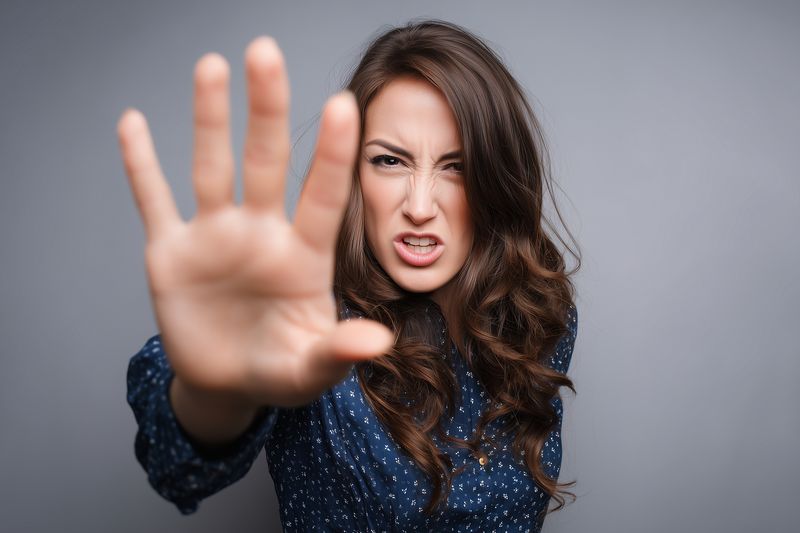 Portrait of a young woman reaching forward with an outstretched hand in a clear stop gesture, showing assertive refusal and intense facial expression against a neutral studio background.