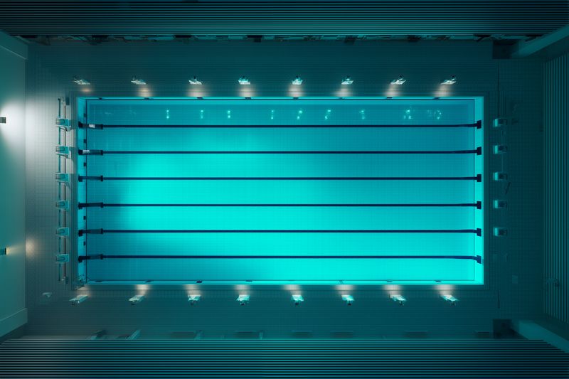 Aerial view of an empty modern indoor swimming pool with illuminated blue water and parallel lanes. Clean tiled deck, subtle reflections and balanced lighting create a calm, symmetrical composition.