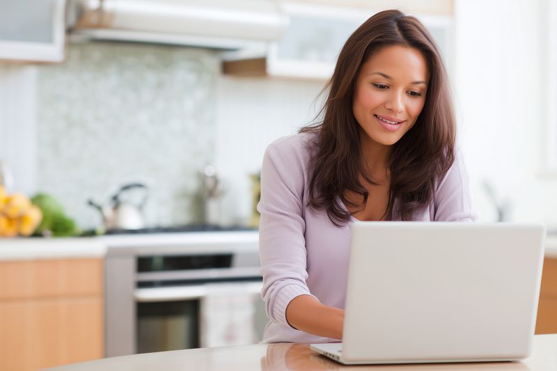 Smiling woman using a laptop at a kitchen counter, working from home and multitasking with a relaxed posture, natural light and cozy domestic interior setting with modern appliance backdrop.