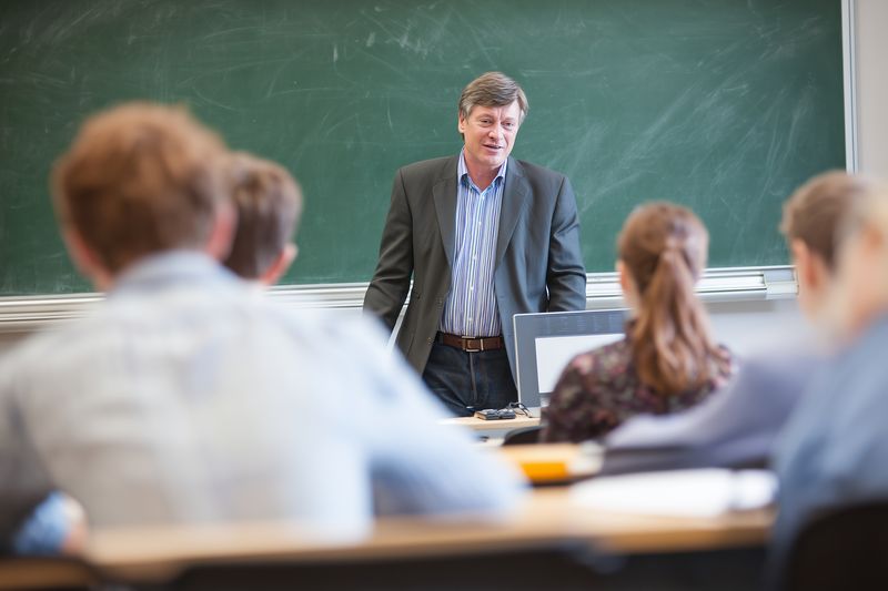 Middle aged male teacher giving a lecture to attentive students in a classroom with a chalkboard behind him, capturing a candid educational moment and interaction during a daytime lesson.